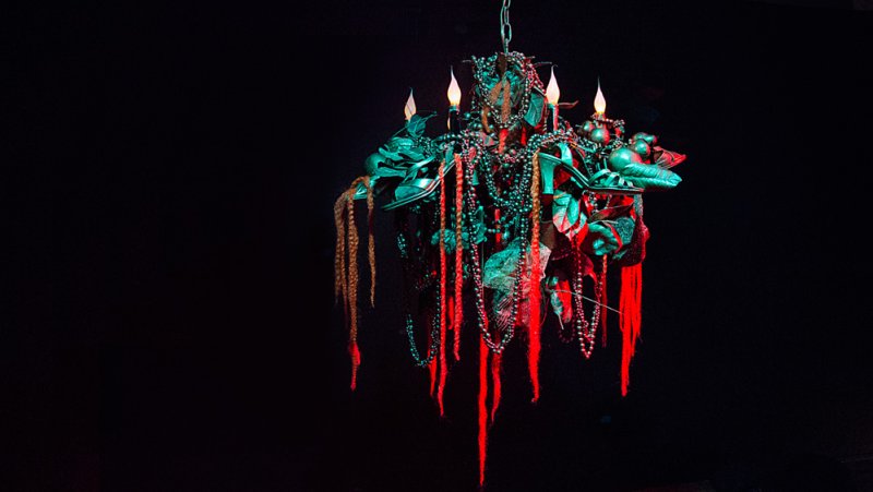 Chandelier with red and green lighting, adorned with beads, leaves, and hanging strands, against a dark background.