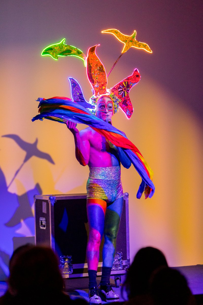 Drag performer Darrell Thorne in a colorful costume with neon bird headdress and rainbow fabric, on stage.