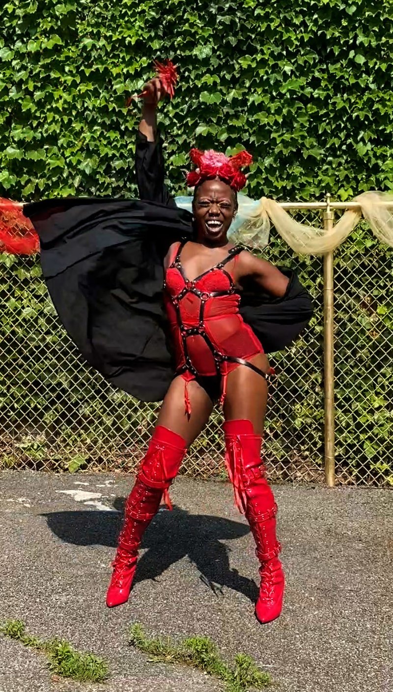 Performer in red lingerie and thigh-high boots, black cape, and elaborate red headpiece, striking a pose in front of a chain-link fence.