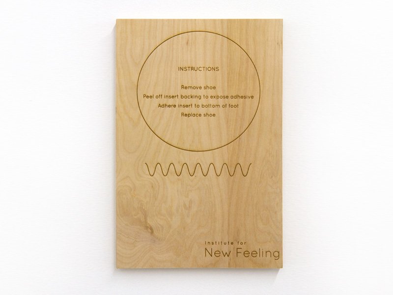 Light wood panel with engraved "INSTRUCTIONS" in a circle, a wavy line, and "Institute for New Feeling" at the bottom.