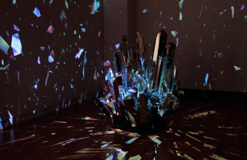 Sculpture of jagged, clear crystals with blue and purple light emanating from within, casting colorful, fragmented reflection
