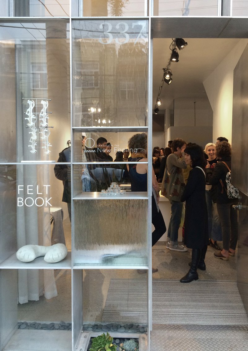A storefront window display with "FELT BOOK" text, two white felt sculptures, and a small succulent garden. Inside, people mingle.