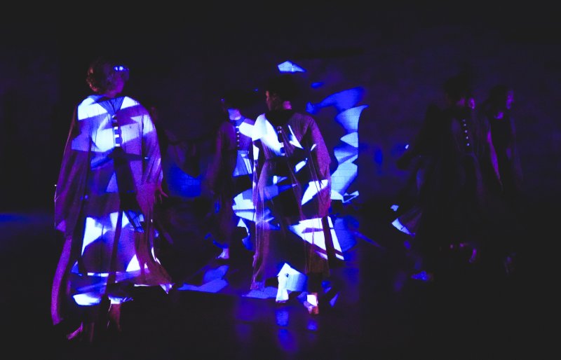 Dancers in dark clothing, illuminated by bright blue and purple geometric projections, move across a dark stage.