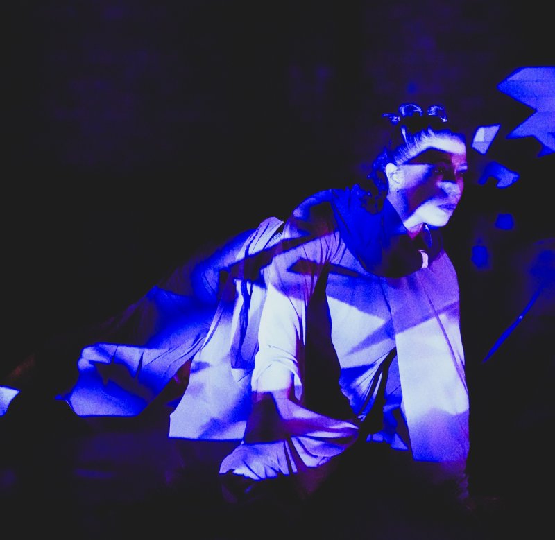 Dancer in a white robe, kneeling on a dark stage, illuminated by blue and purple light with sharp, geometric projections.
