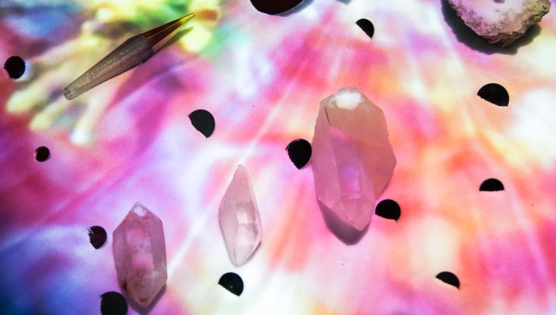 Three pink crystals and a glittery pen on a tie-dye background of pink, purple, yellow, and blue with black half-circles.