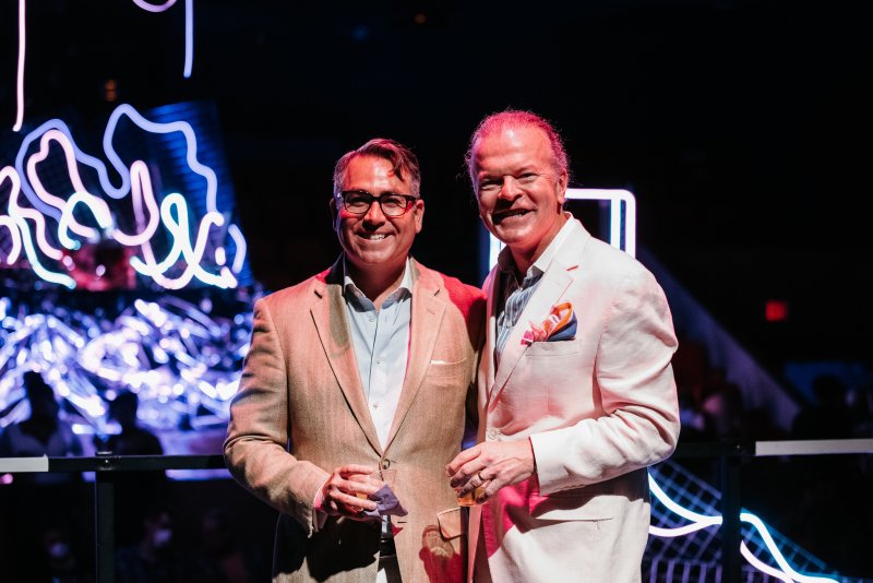 Two men in suits smiling, holding drinks, against a dark background with bright pink, blue, and white neon light art.