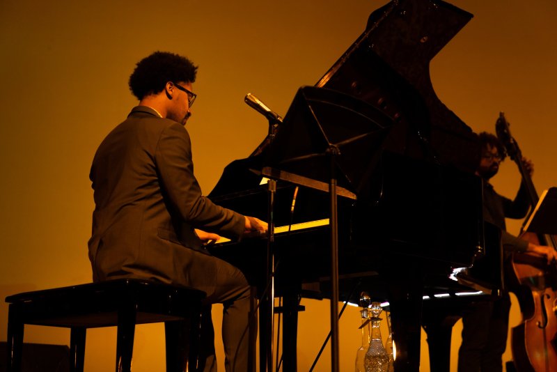 A man in a suit plays a grand piano on a stage bathed in warm, orange light, with a bassist in the background.
