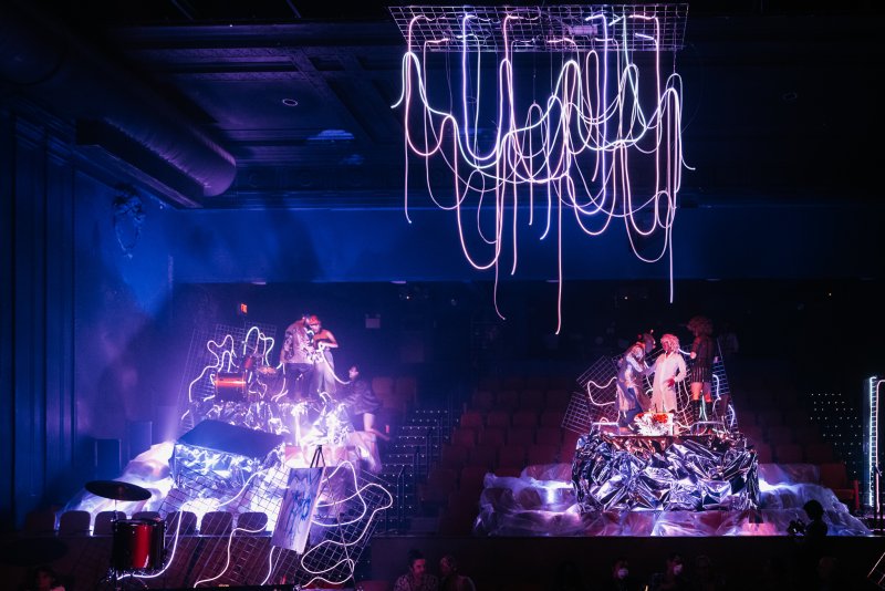 Stage with two glowing platforms, performers, and abstract neon light sculptures hanging above and on the platforms.