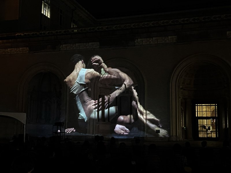 Projection of two intertwined figures, one in white, on a dark building facade with arched windows, viewed by an audience.