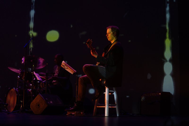 Scott N Andrew's "Suite Life" (2022-2023) features a musician playing guitar on a stool, with a drummer in the background, against a dark stage with projected abstract lights.