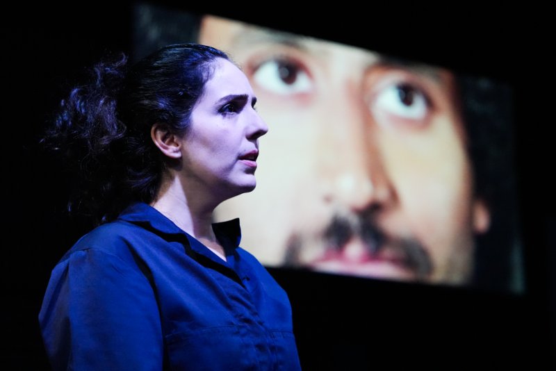 Woman in dark blue shirt in profile, with a man's face projected large behind her looking up and right.