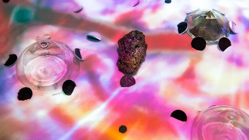 A dark, textured rock sits center on a vibrant pink, purple, and orange surface, surrounded by glass domes and black fragment
