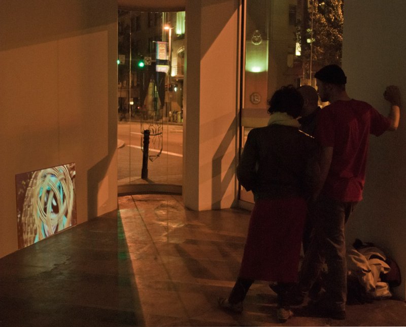 Three people stand in a dimly lit corner next to a window, looking at a small projected abstract image on the wall.