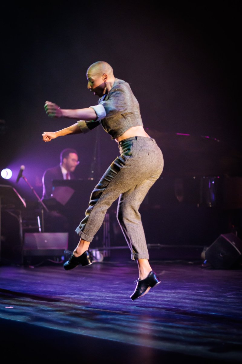 Bald dancer in gray suit mid-leap on a dark stage, with a pianist in the blurred background.