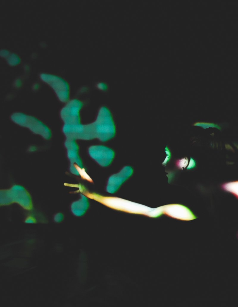 Dancer in profile, reaching towards green abstract light patterns on a dark wall.