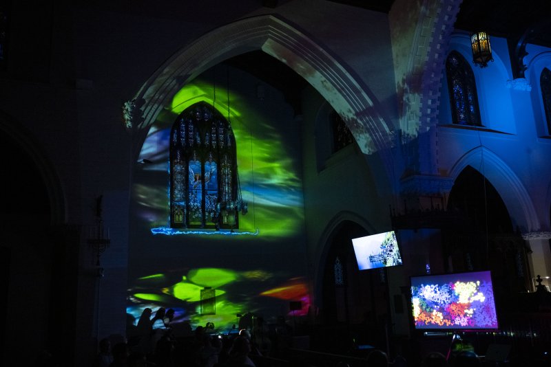 Green and blue light projections illuminate a dark church interior, highlighting a stained-glass window and two screens displaying abstract art.