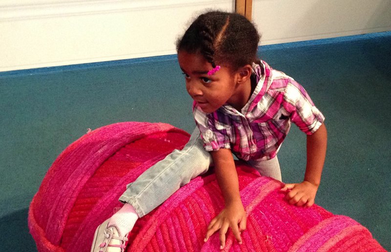 Young Black girl in a pink plaid shirt and jeans on a large, textured pink sculpture, looking left.
