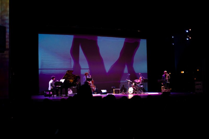 Jazz quartet on a dark stage, large screen behind them displaying a purple-hued abstract image.