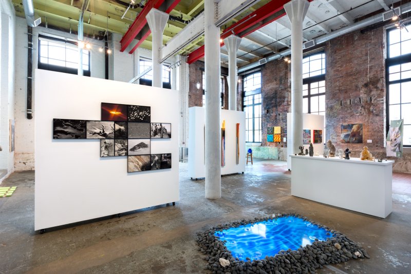 Art exhibition in a large industrial space with a glowing blue "reflecting pool" surrounded by dark rocks.