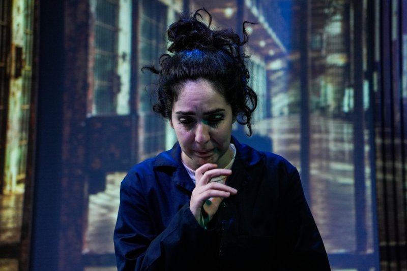 Woman with messy dark hair and smudged makeup, wearing a dark blue jumpsuit, looks down with a pained expression against a blurred urban background.