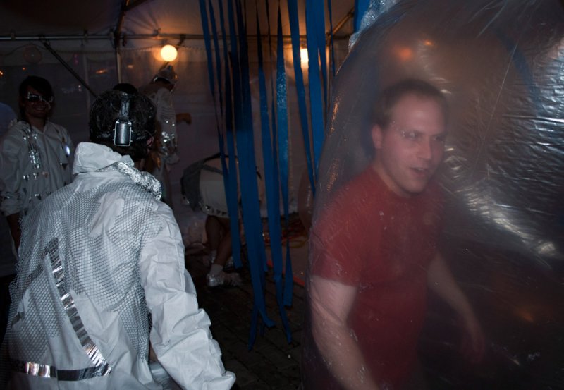 Man in red shirt inside clear plastic bubble, surrounded by people in white suits and blue streamers.