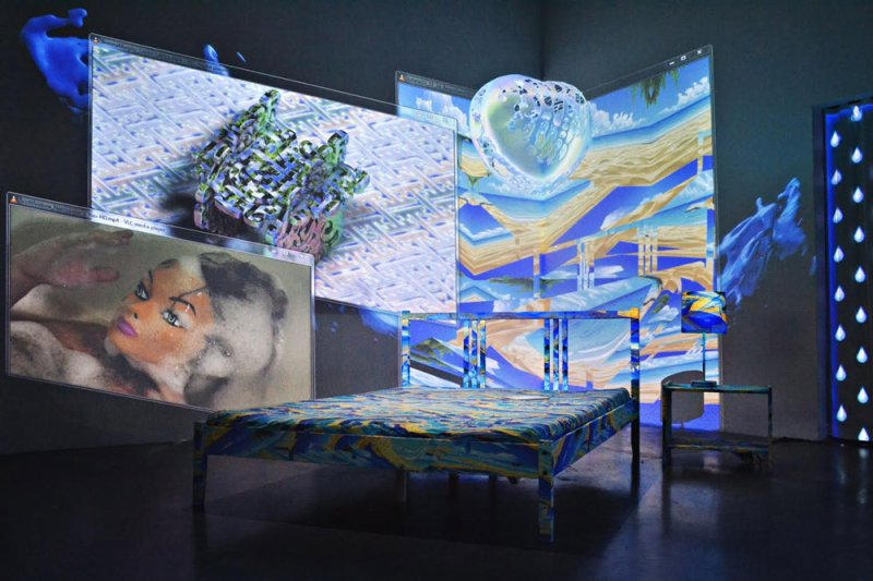 Installation with a bed, screen projections of abstract landscapes, a doll, and a heart, all in blues and yellows.