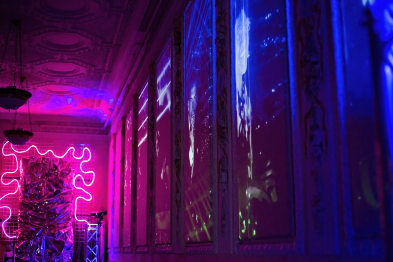 Neon pink squiggly light sculpture and blue/purple projected figures on ornate white panels in a dimly lit room.