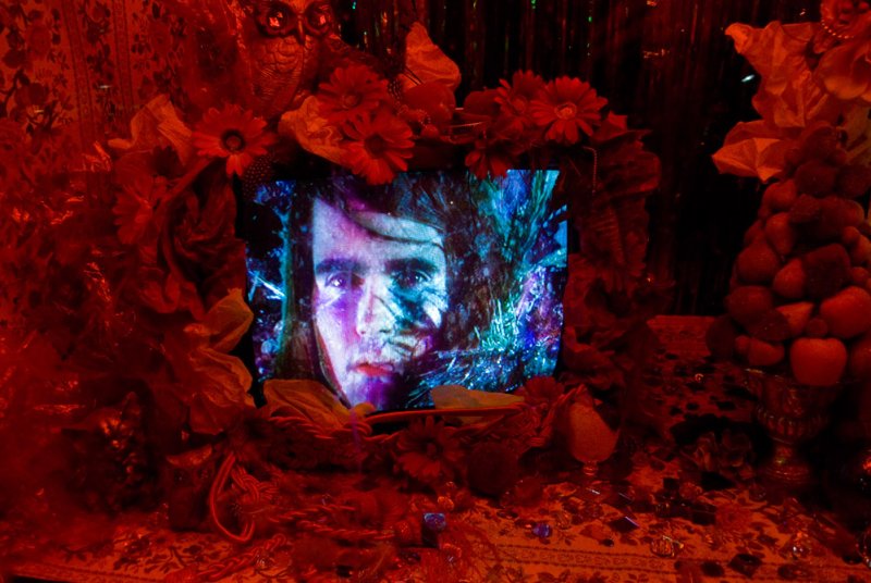 Artwork with a glowing screen displaying a man's face in blue and purple hues, surrounded by red flowers and fabrics.