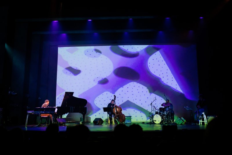 Scott N Andrew's "Suite Life" (2022-2023) shows four musicians on stage, with a large screen displaying abstract purple and white shapes.