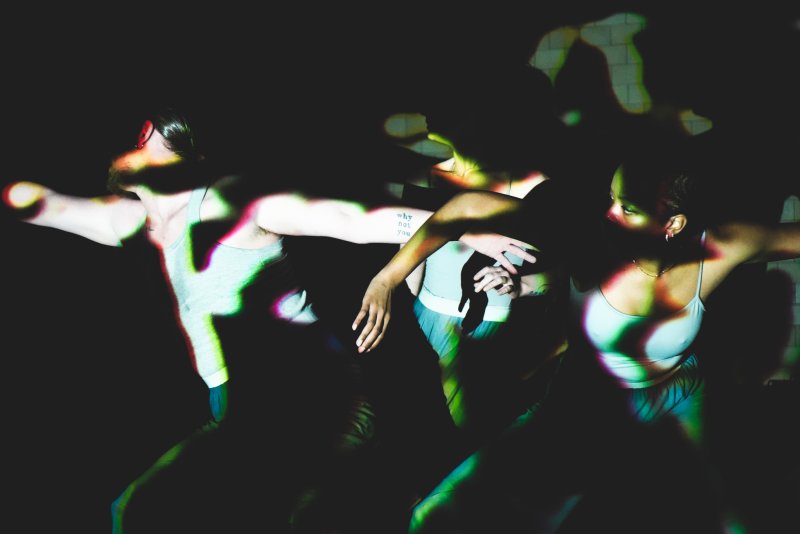 Three dancers in motion, illuminated by green and purple light on a dark background.