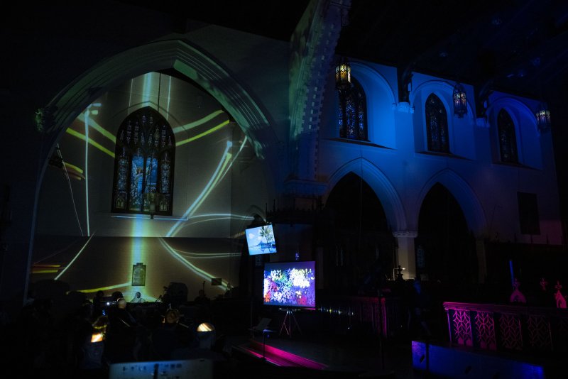 Dark church interior with green and blue light projections, stained glass, and two screens displaying abstract art.