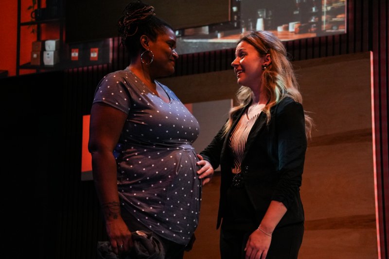 Two women, one in a polka dot top and the other in a black blazer, smiling and looking at each other in a dimly lit room.