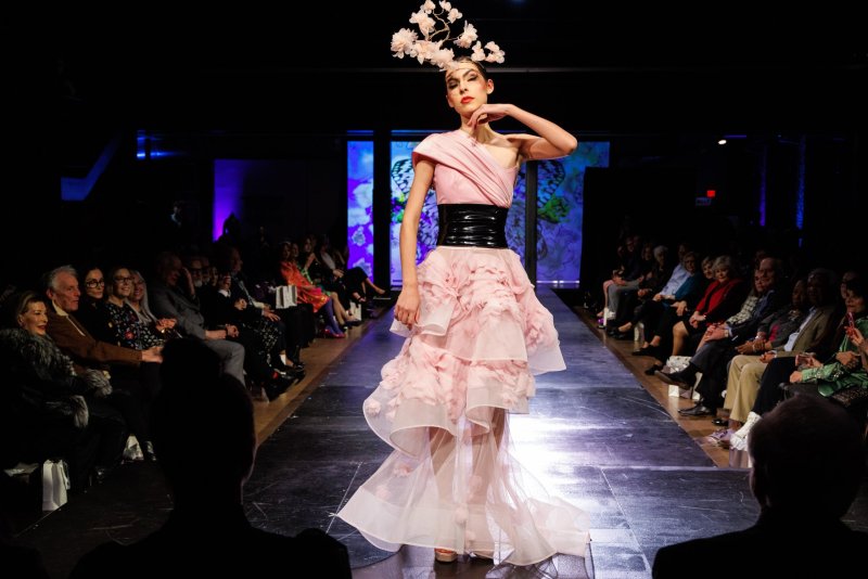 Fashion model in a pink ruffled gown with a black belt and floral headpiece walks a runway before a seated audience.