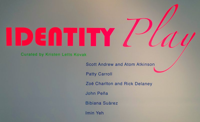 Exhibition title "IDENTITY Play" in large pink script, with artist names listed below in blue on a gray wall.