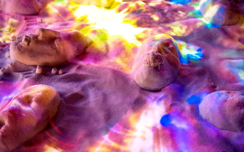 Sculpted faces and small objects embedded in sand, illuminated by shifting pink, purple, blue, and yellow light.