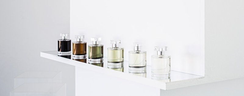 Six perfume bottles with clear caps, varying in liquid color from dark brown to light yellow, on a reflective white shelf.