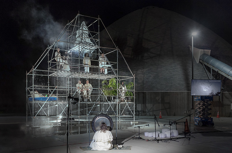 Performance art with people on a scaffolding structure under falling water, a gong, and a screen, against a dark, pyramid-like building.