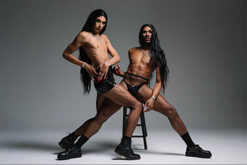 Two drag queens with long black hair, one in a black vinyl skirt and fishnets, the other in black briefs and fishnets, posing against a gray background.